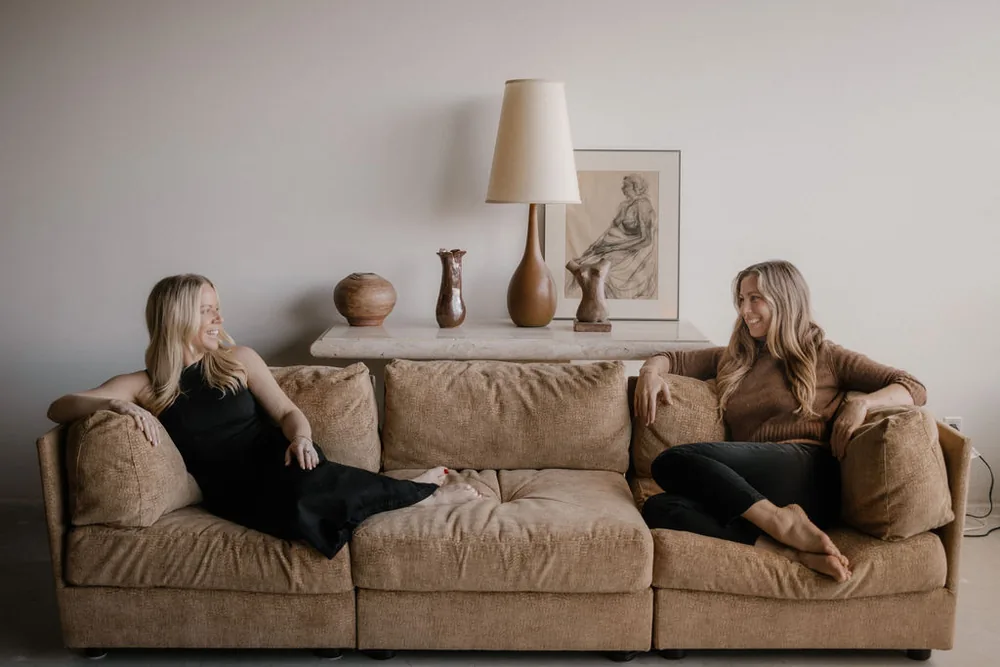 Emily and Cait sitting at either end of a long sofa, smiling at each other