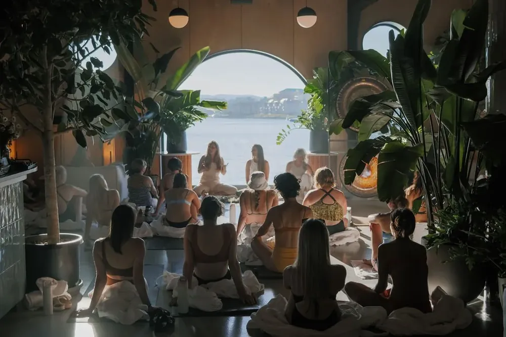 A circle of women seated on the floor of a room with an arched window onto the water